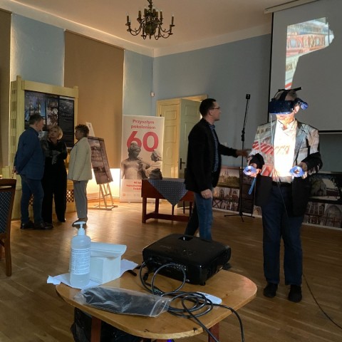 Powiększ obraz: Uczestnik konferencji w goglach wirtualnej rzeczywistości i kontrolerami sterującymi zwiedza wirtualny zabytek.